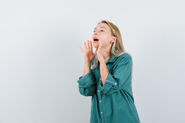 Chica rubia en blusa verde cogidos de la mano cerca de la boca como llamando a alguien y mirando enfocado