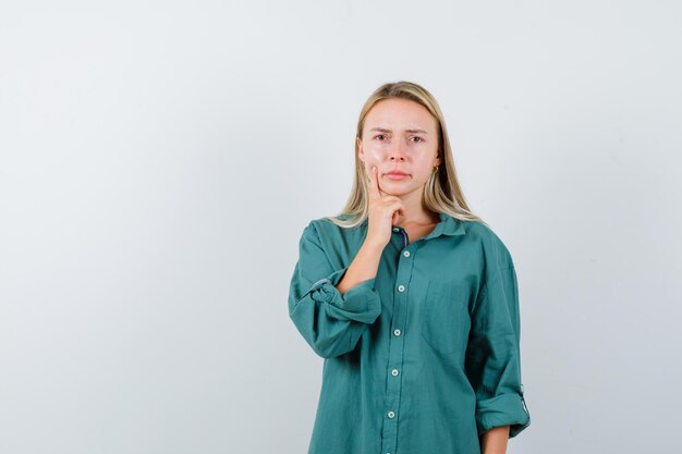 Chica rubia en blusa verde poniendo el dedo índice cerca de la boca, pensando en algo y mirando pensativo