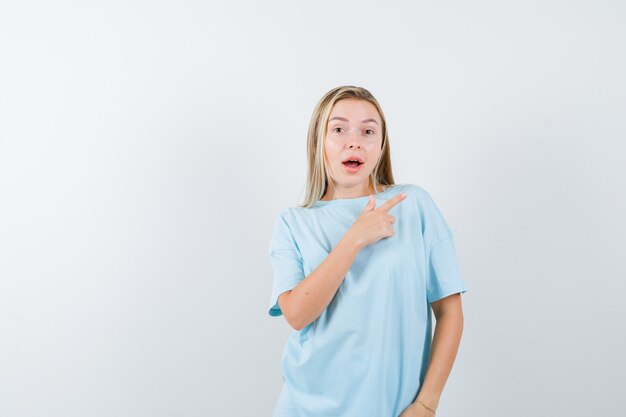 Chica rubia en camiseta azul apuntando a la derecha con el dedo índice y mirando sorprendido, vista frontal.