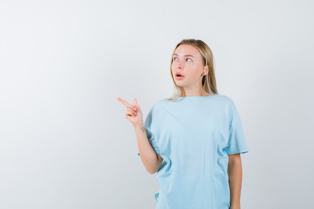 Chica rubia en camiseta azul apuntando hacia la izquierda con el dedo índice y mirando enfocado, vista frontal.