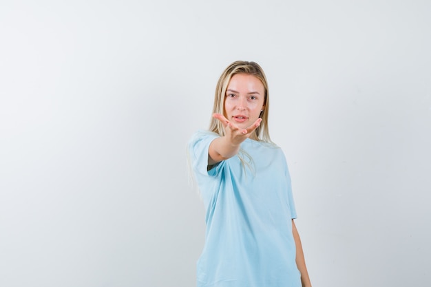 Chica rubia en camiseta azul estirando la mano hacia la cámara mientras sostiene algo y mira seria, vista frontal.