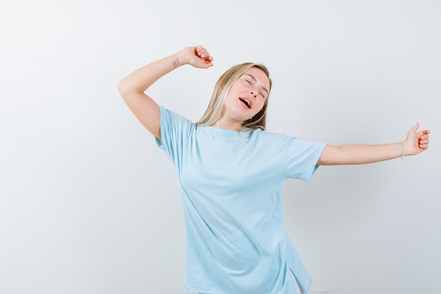 Chica rubia en camiseta azul estirándose y bostezando, manteniendo los ojos cerrados y mirando con sueño, vista frontal.