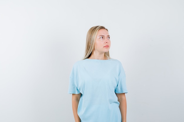 Chica rubia en camiseta azul mirando a otro lado mientras posa a la cámara y se ve bonita, vista frontal.