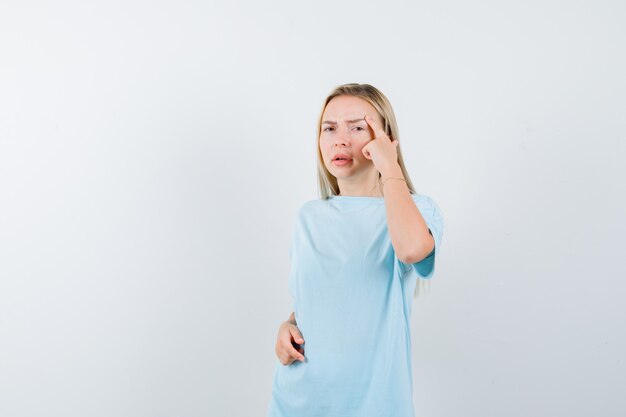 Chica rubia en camiseta azul poniendo el dedo índice en la sien y mirando pensativo, vista frontal.