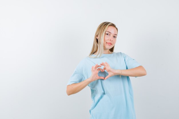 Chica rubia en camiseta azul que muestra el gesto del corazón y se ve bonita, vista frontal.