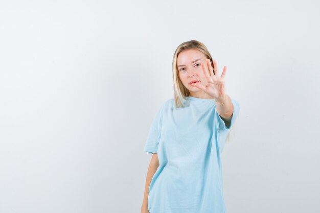 Chica rubia en camiseta azul que muestra la señal de stop y mirando seria, vista frontal.