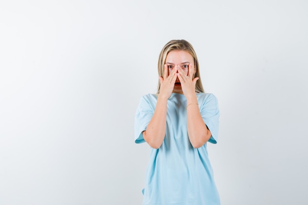 Chica rubia en camiseta azul que muestra signos v en los ojos, mirando a través de los dedos y mirando sorprendido, vista frontal.