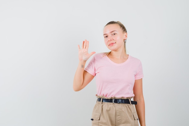 Chica rubia en camiseta, pantalones agitando la mano para decir adiós y mirando alegre, vista frontal.