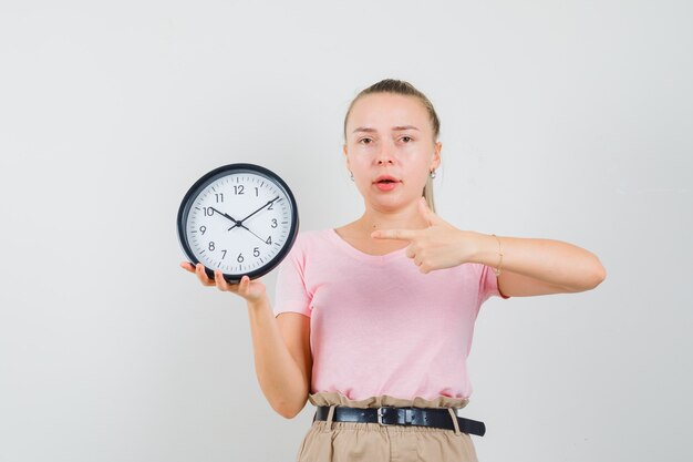 Chica rubia en camiseta, pantalones apuntando al reloj de pared y mirando sensible, vista frontal.