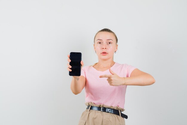 Chica rubia en camiseta, pantalones apuntando al teléfono móvil y mirando perplejo, vista frontal.