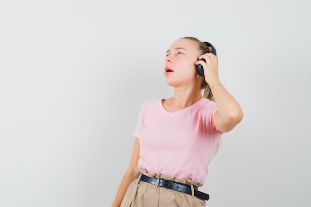 Chica rubia en camiseta, pantalones escuchando música con auriculares y mirando encantada, vista frontal.