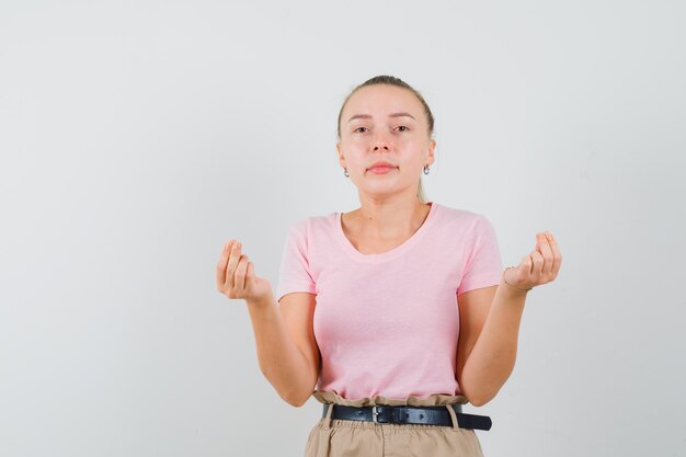 Chica rubia en camiseta, pantalones haciendo gesto de dinero, vista frontal.