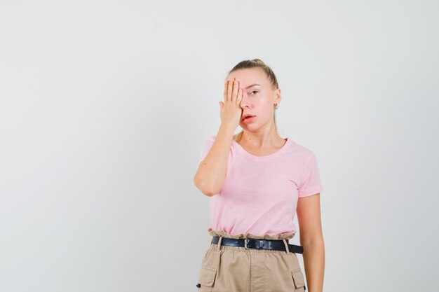 Chica rubia en camiseta, pantalones sosteniendo la mano en un ojo y luciendo elegante, vista frontal.