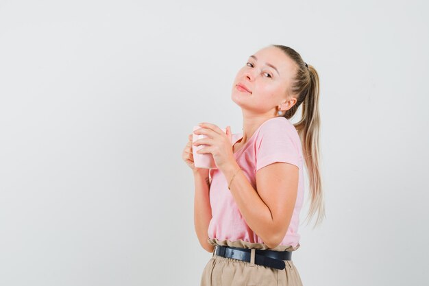 Chica rubia en camiseta, pantalones sosteniendo una taza de bebida y luciendo bonita, vista frontal.