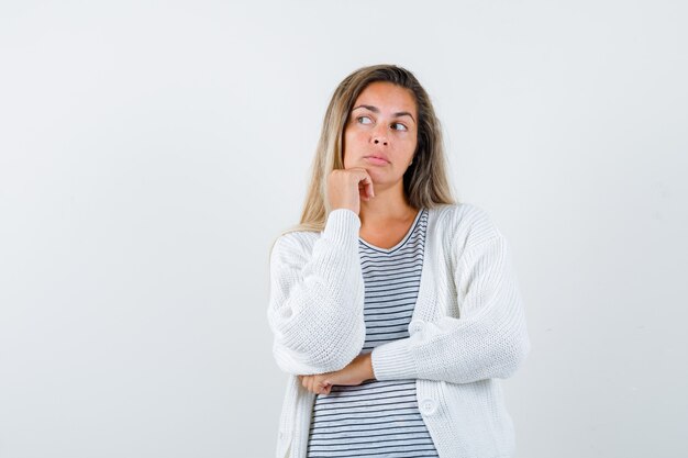 Chica rubia con camiseta a rayas, cárdigan blanco y pantalón de mezclilla apoyando la barbilla en la mano mientras sostiene la mano debajo del codo y mira pensativa, vista frontal.