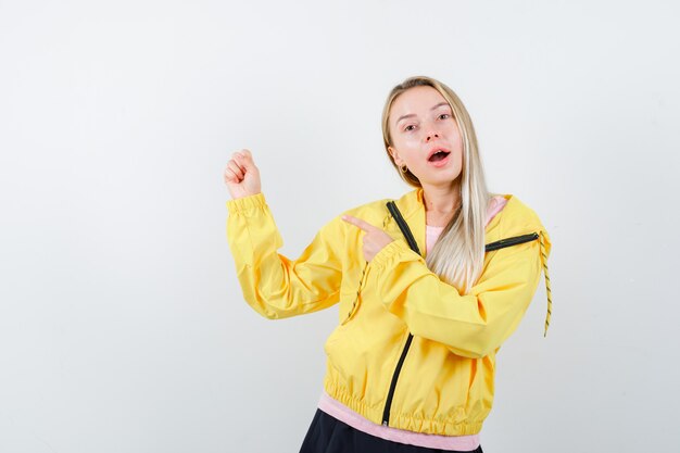 Chica rubia en camiseta rosa y chaqueta amarilla apretando el puño y señalando y mirando feliz