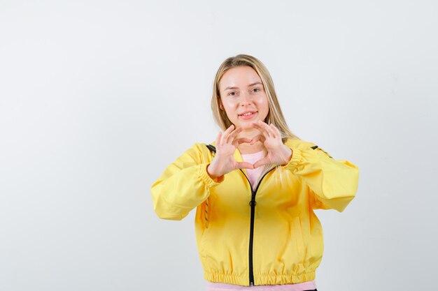 Chica rubia en camiseta rosa y chaqueta amarilla mostrando gesto de amor con las manos y mirando seductor