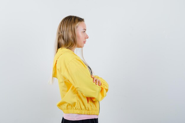 Chica rubia en camiseta rosa y chaqueta amarilla de pie con los brazos cruzados y mirando serio