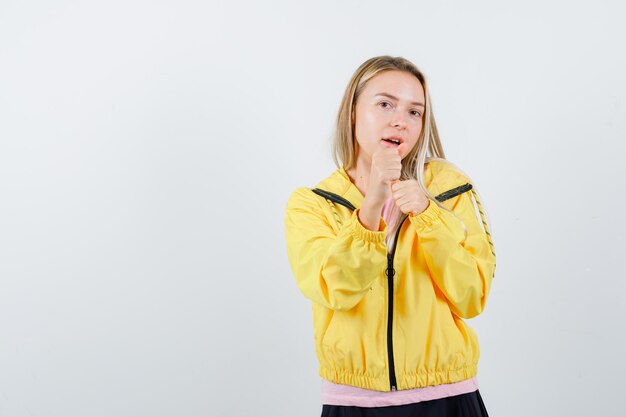 Chica rubia en camiseta rosa y chaqueta amarilla de pie en pose de boxeador y mirando poderosa