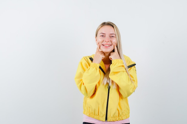 Chica rubia con camiseta rosa y chaqueta amarilla sosteniendo los dedos índices cerca de la boca, forzando una sonrisa y luciendo feliz