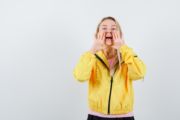 Chica rubia en camiseta rosa y chaqueta amarilla tomados de la mano cerca de la boca como llamando a alguien y mirando enfocado