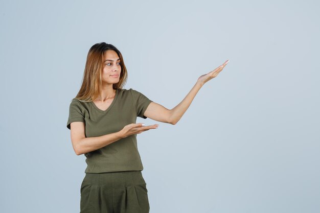 Chica rubia en camiseta verde oliva y pantalones estirando las manos como sosteniendo y presentando algo, mirándolo y mirando enfocado, vista frontal.