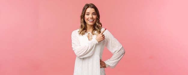 Chica rubia caucásica de buen aspecto optimista con vestido blanco apuntando a la esquina superior derecha y sonriendo a la cámara con cara curiosa interesada discutir promociones especiales descuentos de primavera fondo rosa