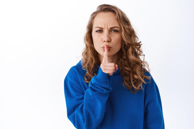 Una chica rubia con el ceño fruncido enojada calla y dice que se calle pidiendo silencio haciendo un gesto de silencio presionando el dedo en los labios fondo blanco