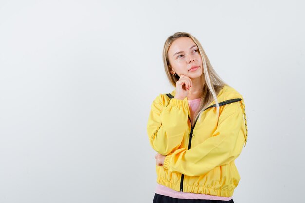 Chica rubia en chaqueta amarilla apoyando la barbilla en la mano y mirando pensativa