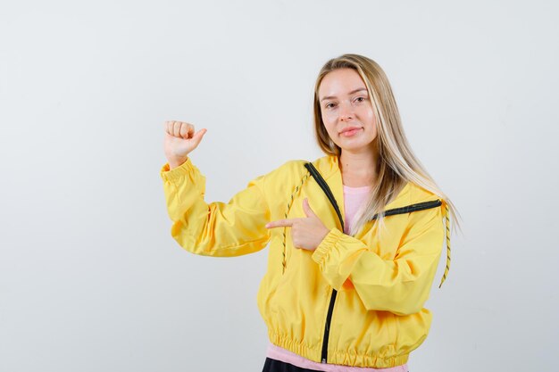 Chica rubia en chaqueta amarilla apuntando a un lado mientras posa y mira alegre