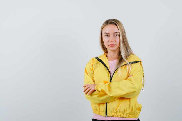 Chica rubia en chaqueta amarilla de pie con los brazos cruzados y mirando confiada