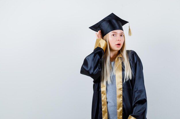 Chica rubia cogidos de la mano cerca de la oreja para escuchar en toga y gorra de graduación y mirando sorprendido.