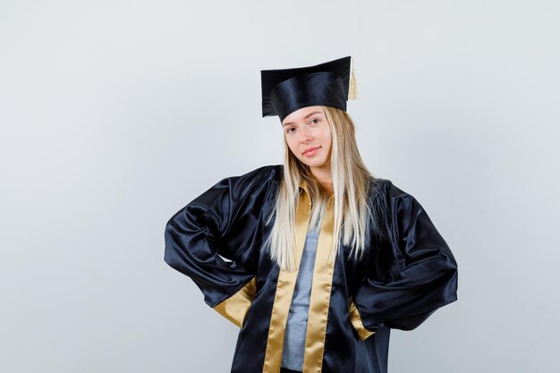 Chica rubia cogidos de la mano en la cintura en toga y gorra de graduación y luciendo linda.