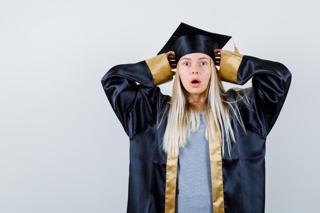 Chica rubia cogidos de la mano en la tapa con la boca abierta en toga y gorra de graduación y mirando sorprendido.