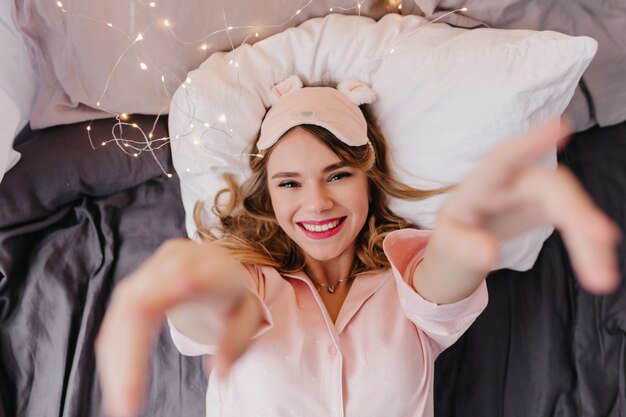 Chica rubia complacida en sábana oscura. Fotografía aérea interior de la sonriente dama alegre en rosa eyemask posando en la cama.