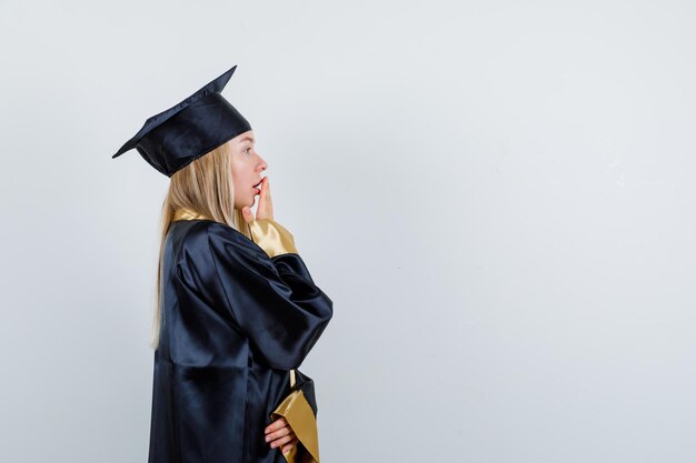 Chica rubia cubriendo la boca con la mano en toga y gorra de graduación y mirando sorprendido