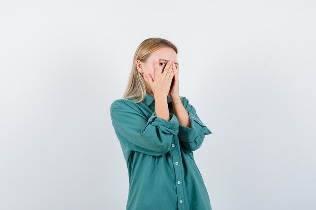 Chica rubia cubriendo la cara con la mano y mirando a través de los dedos en blusa verde y mirando tímida.