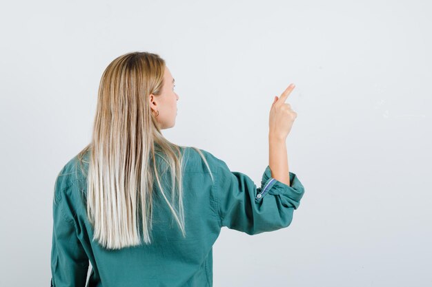 Chica rubia dando vuelta y apuntando a la derecha en blusa verde y con aspecto encantador