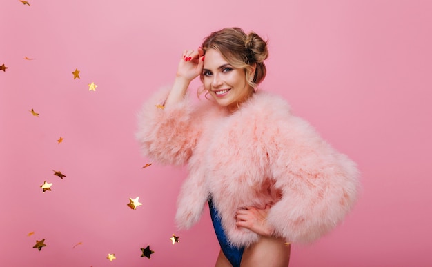 Chica rubia delgada alegre divirtiéndose en la fiesta de cumpleaños posando con confeti dorado sobre fondo rosa. Atractiva mujer joven sonriente en abrigo de piel con estrellas de brillo de pie delante de la pared brillante