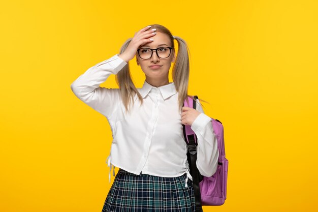 Chica rubia del día mundial del libro tocando la frente con mochila sobre fondo amarillo