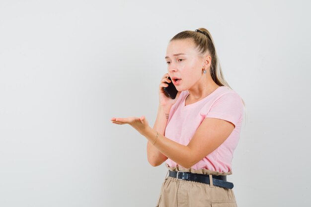 Chica rubia discutiendo algo en el teléfono móvil en camiseta, vista frontal de los pantalones.