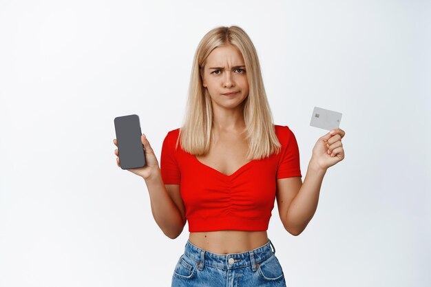 Una chica rubia disgustada hace una mueca y muestra la pantalla del teléfono móvil y la tarjeta de crédito haciendo pucheros molesto de pie sobre fondo blanco.