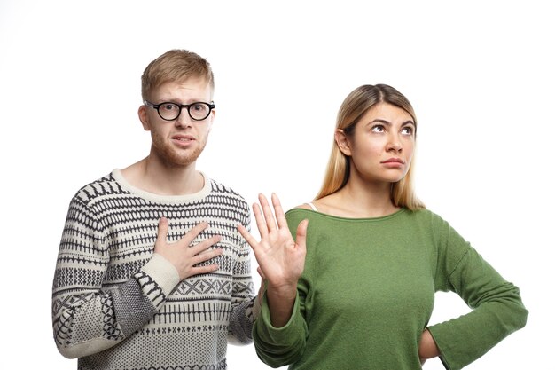 Chica rubia disgustada que muestra la señal de alto con la palma de la mano, lo que significa que se acabó mientras su infeliz y miserable novio con mirada infeliz sosteniendo la mano en su pecho como señal de que se ha roto.