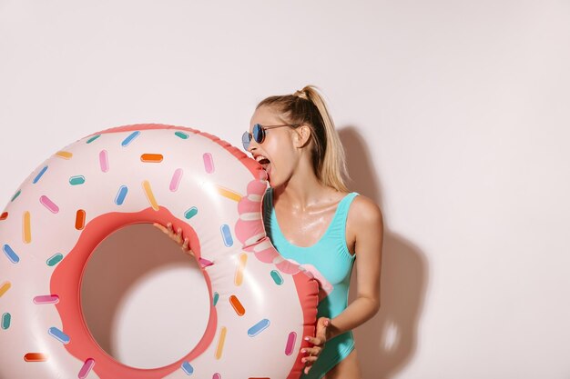 Chica rubia divertida con gafas de sol y traje de baño brillante posando con un gran anillo de natación en forma de rosquilla en un fondo aislado