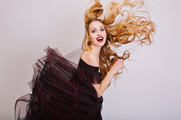 Chica rubia divirtiéndose, hermoso cabello largo y rizado en el aire, mujer joven posando. Mirada divertida con la boca abierta. Con vestido negro con falda esponjosa, maquillaje brillante. Aislado..