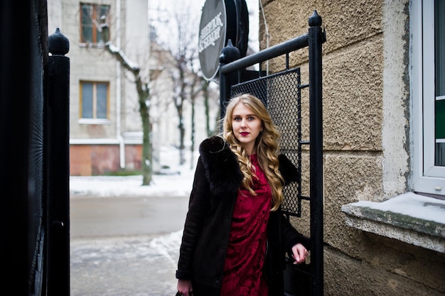 Chica rubia de elegancia en vestido de noche rojo y abrigo de piel en las calles de la ciudad en el día de invierno