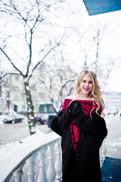 Chica rubia de elegancia en vestido de noche rojo y abrigo de piel en las calles de la ciudad en el día de invierno