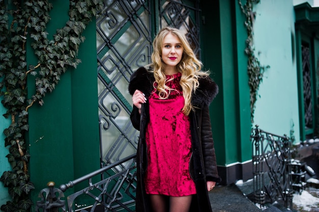 Chica rubia de elegancia en vestido de noche rojo y abrigo de piel en las calles de la ciudad