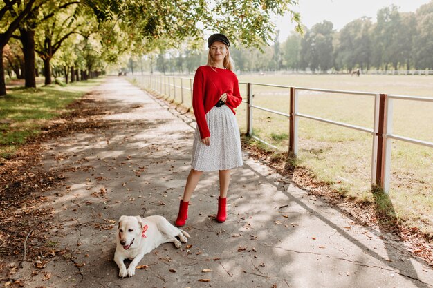 Chica rubia elegante con sombrero negro y falda blanca posando con su perro en el camino del parque.