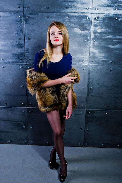 Chica rubia elegante vestida con abrigo de piel y vestido combinado contra la pared de acero en el estudio